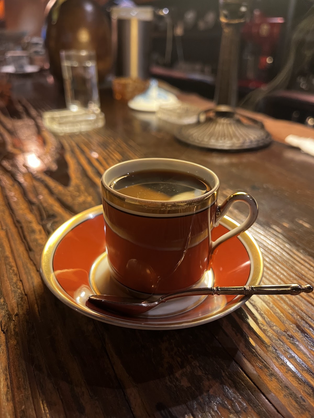 木目のカウンターに赤茶金縁の陶器カップとコーヒー、背後に金属製の小物類が並ぶ純喫茶のカウンター