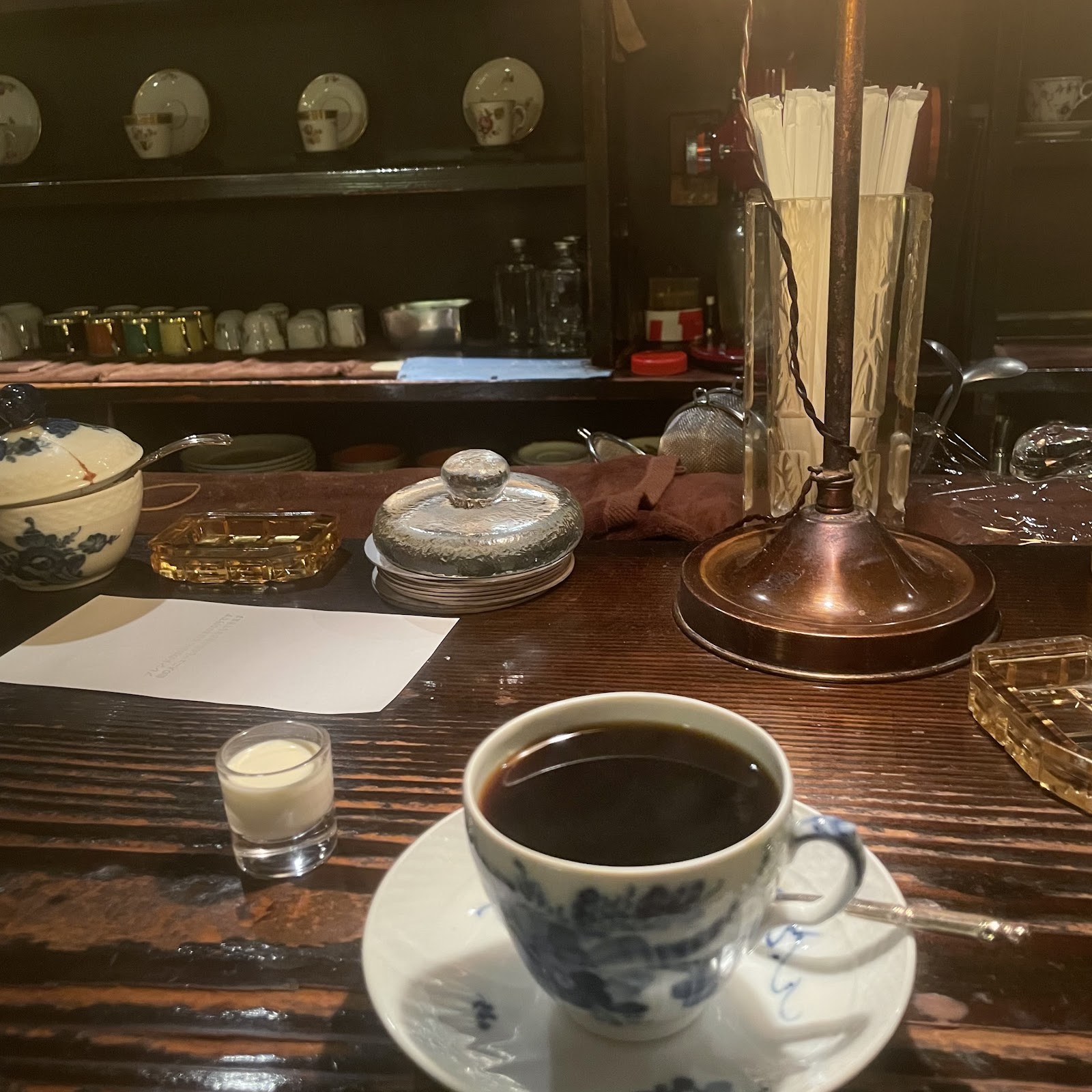 飴色に光る木製カウンターに置かれた染付け柄の白磁カップとコーヒー、奥の棚にカップ類が整然と並ぶ純喫茶の内観