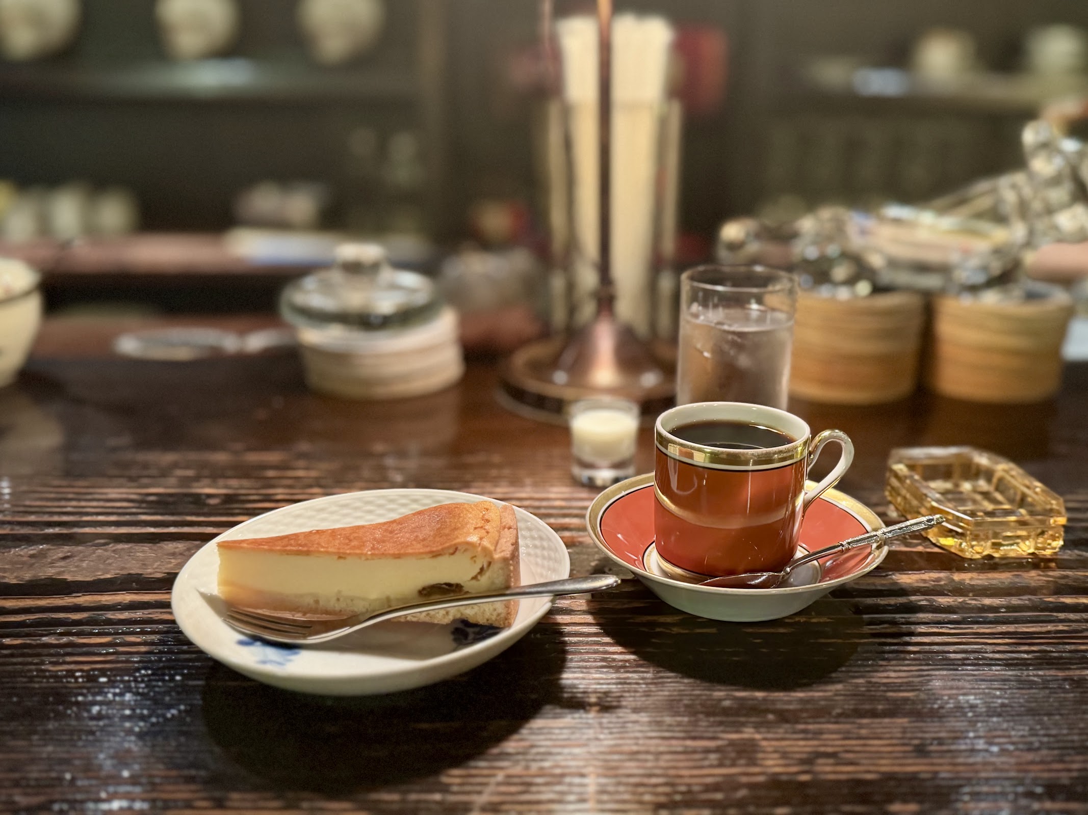 木製カウンターの上に赤茶色の金縁カップに注がれたコーヒーと白皿のチーズケーキ、奥にガラス食器が並ぶ純喫茶の卓上